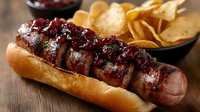 Grilled sausage in toasted bun topped with cranberry sauce and fresh herbs, served with crispy potato chips on rustic wooden table, close up view.