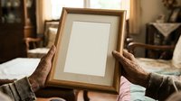 Senior man with dark skin tone holding empty wooden frame with white mat in home interior, displaying artwork or family photo.