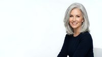 Mature woman with long gray hair and fair complexion wearing black top, smiling at camera against white background for portrait and lifestyle use.