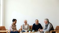 Four mature adults of diverse ethnicities engaged in casual conversation around a wooden table with beverages and plants, representing friendship, community gathering, and social connection for lifestyle and editorial use.