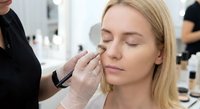 Professional makeup artist applying eyeshadow to young blonde woman client in modern beauty salon with cosmetic products in background.