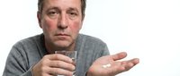 Middle-aged Caucasian man holding pills and glass of water, appearing concerned or hesitant about medication. Studio portrait on white background.