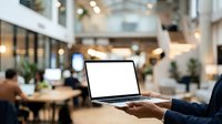 Businessperson holding laptop with blank screen in modern office lobby or coworking space with blurred background, suitable for corporate technology and business concepts.