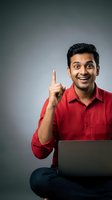 Excited young Indian businessman in red shirt pointing finger up with eureka expression while working on laptop against gray studio background.