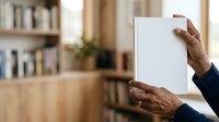 Senior man with dark skin tone holding blank white card or paper in hands, displaying mockup space for text or design, indoor home library setting with bookshelves.