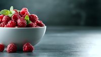 Fresh red raspberries in white bowl with mint leaves on dark wooden surface for healthy eating and organic food concepts.