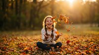 Young blonde girl sitting cross-legged in autumn park throwing colorful fall leaves in air with warm golden sunlight creating magical atmosphere.