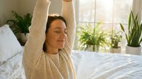 Relaxed woman with dark hair and olive complexion stretching arms overhead on bed in bright bedroom with potted plants and natural sunlight, conveying wellness and morning routine.