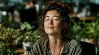 Young woman with curly brown hair relaxing with eyes closed in modern cafe or restaurant setting with natural lighting and green plants in background.