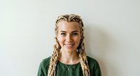 Young woman with blonde braided hair and fair complexion wearing a green shirt, smiling at camera against white background. Portrait for lifestyle, fashion, and beauty content.