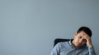 Young adult male with dark hair and medium complexion wearing casual light blue shirt, resting head on hand against neutral gray background, expressing stress, fatigue, or contemplation for business, mental health, or lifestyle concepts.