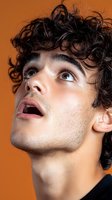 Young Mediterranean male model with curly dark hair looking up expressively against orange background, showcasing dramatic facial features and natural makeup styling.