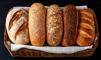 Fresh artisanal bread loaves with various seeds and toppings arranged in wicker basket on white cloth. Rustic bakery display shows different types of crusty bread.