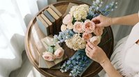 Woman arranging pastel pink roses and blue hydrangea flowers in round mirror tray for wedding decor and floral design projects.