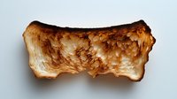 Burnt toast slice with uneven charred surface and crispy texture isolated on white background, showing detailed toasted bread structure and browning pattern.