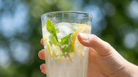 Hand holding refreshing mojito cocktail with fresh mint leaves, lime, and ice against blurred outdoor garden background, perfect for beverage and summer drink concepts.