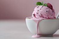 Melting raspberry ice cream scoop in white ceramic bowl garnished with fresh berries and mint leaves, pink drip flowing down side on light background.