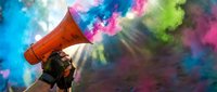 Hand holding megaphone releasing vibrant multicolored powder smoke in pink, blue and green hues against a sunlit forest backdrop.