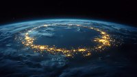 Aerial view of Earth at night showing illuminated coastal cities forming glowing ring around ocean, captured from space with curved horizon and dark atmosphere.