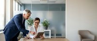 Mature male executive in navy suit and middle-aged female professional in white blouse reviewing digital tablet together at wooden desk in modern office with glass partitions and plants.