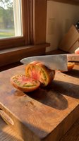 Ripe tomato sliced in half with chef's knife on wooden cutting board near window with natural sunlight, suitable for cooking, recipe, and food preparation content.