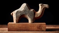 Ancient stone camel figurine on wooden base against dark background, showcasing primitive art sculpture with minimalist design and natural textures.