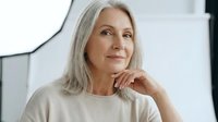 Mature woman with long gray hair and fair complexion, wearing casual beige top, hand near face in thoughtful pose, indoor setting with white background.