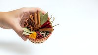 Hand holding wicker basket filled with aromatic spices including cinnamon sticks, star anise, bay leaves, peppercorns and turmeric on white background.