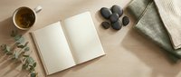 Cozy flat lay with open blank notebook, herbal tea cup, spa stones and eucalyptus on wooden surface for wellness journaling and mindful relaxation.