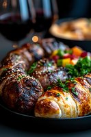 Grilled meat medallions and golden baked croissant served with fresh herbs and colorful vegetables on dark plate, red wine in background, moody restaurant lighting.