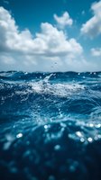 Dynamic ocean wave splashing against deep blue waters under white fluffy clouds and bright sky, creating sparkling water droplets and bokeh effects.