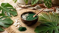 Spirulina powder in a coconut bowl with a wooden spoon, surrounded by tropical leaves and coconut flakes on a woven mat.