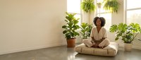 Woman with curly hair, medium skin tone, sitting cross-legged on floor surrounded by potted plants near bright window in minimalist room for wellness and mindfulness content.