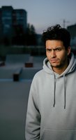 Young man with dark wavy hair and facial hair, Middle Eastern ethnicity, wearing gray hoodie, standing outdoors against urban cityscape backdrop with blurred buildings and water.
