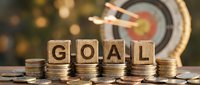 Business goal achievement concept with wooden letter blocks spelling GOAL on stacked coins with dartboard and arrow hitting bullseye target in background.