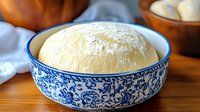 Fresh homemade yeast dough rising in traditional blue and white ceramic bowl, covered in flour, ready for baking bread or pastries on wooden surface.