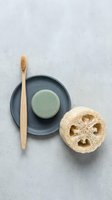 Natural spa essentials with bamboo toothbrush, green soap bar on ceramic plate and loofah sponge arranged on concrete surface for wellness routines.