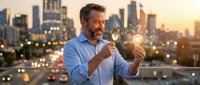 Middle-aged Caucasian businessman examining light bulb with magnifying glass on city rooftop at sunset, symbolizing innovation and business analysis.
