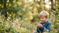 Young boy with blonde hair wearing denim shirt exploring nature outdoors, picking wildflowers in green garden setting for lifestyle and childhood themes.