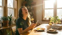 Woman with long dark hair and medium skin tone sitting at wooden table by sunlit windows, holding smartphone, surrounded by potted plants, food plates, and books, enjoying morning breakfast in bright home interior.