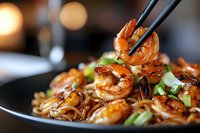 Grilled teriyaki shrimp with noodles on black plate, chopsticks picking up glazed jumbo shrimp. Garnished with fresh green onions, close-up shot with bokeh background.