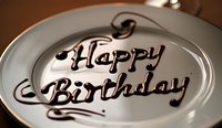 Happy Birthday message written in chocolate syrup on white plate with elegant swirl decorations, close-up view with shallow depth of field for festive mood.