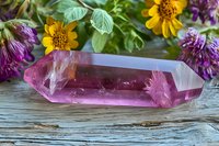 Large amethyst crystal point on rustic wooden surface surrounded by fresh yellow daisies and purple clover flowers, displaying natural healing properties.