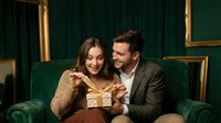 Young couple sharing gift box on luxury green velvet sofa in elegant interior with golden frames for holiday celebration and romantic moments.