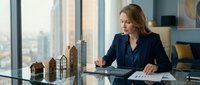 Caucasian female architect or real estate professional in dark blazer reviewing building models and blueprints at modern office desk with city skyline view through windows.