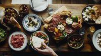 Mediterranean feast spread on rustic wooden table with various appetizers, cured meats, cheeses, olives, and hands reaching for food, captured from above.