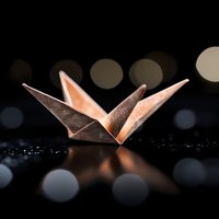 Glowing copper origami butterfly on dark reflective surface with bokeh lights in background creates magical atmospheric composition.