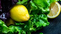 Fresh lemon and vibrant green lettuce leaves with water droplets on dark wooden surface for healthy cooking and salad preparation concepts.