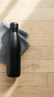Black insulated water bottle with condensation droplets on gray fabric napkin, placed on light wooden floor with natural sunlight casting shadows. For product mockups.