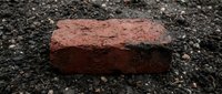 Single weathered red clay brick lying on rough asphalt surface. Construction material for building projects, renovation work, and architectural design.